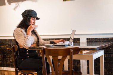 Latin moda tasarımcısı bir kafeteryada dizüstü bilgisayarıyla bazı tasarımlar yapıyor, kahve içiyor.