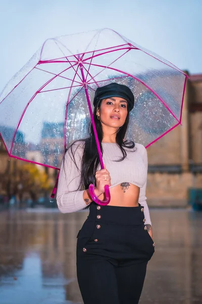 一个年轻的拉丁黑发姑娘带着一顶皮帽站在城市的雨中 带着一把透明的雨伞 — 图库照片
