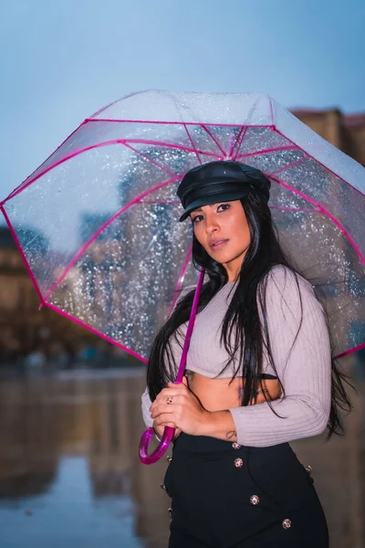 一个年轻的拉丁黑发姑娘在雨中戴着皮帽 带着一把透明的雨伞 — 图库照片