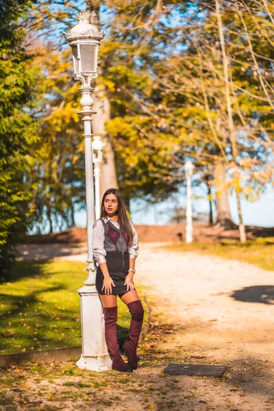 Lifestyle, a young Caucasian brunette college girl, in a park next to a white lamppost enjoying the fall
