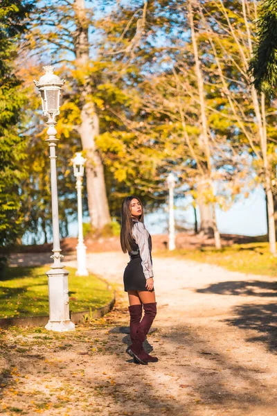 Lifestyle, a young Caucasian brunette college girl, in a park next to a white lamppost enjoying the fall