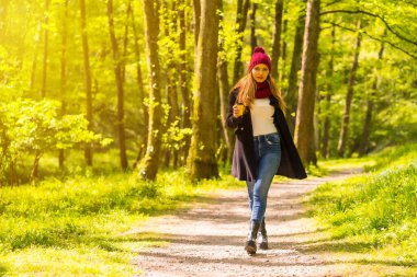 Siyah ceketli, eşarplı ve kırmızı yün şapkalı Latin kız sonbahar parkında eğleniyor, gün batımında bir kahve eşliğinde yolda yürüyor.