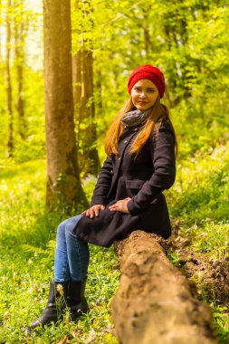 Siyah ceketli ve kırmızı şapkalı bir kadın sonbahar parkında gün batımında gülümsüyor.