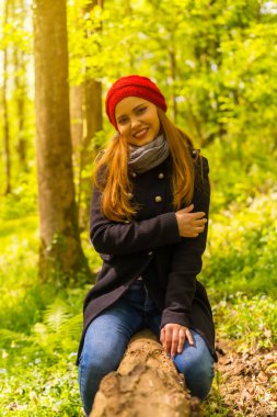 Siyah ceketli ve kırmızı şapkalı bir kadın sonbahar parkında eğleniyor, gün batımında gülümseyen bir ağaçta oturuyor.