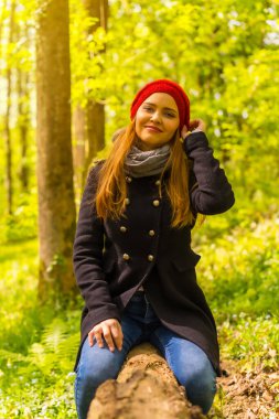 Siyah ceketli ve kırmızı şapkalı bir kadın sonbahar parkında gün batımında bir ağaçta oturmuş gülümsüyor.