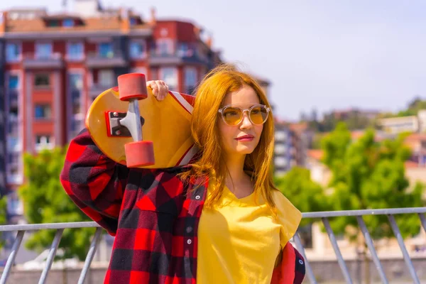 身穿黄色衬衫 红色格子花衬衫和太阳镜的滑板女子 姿势与滑板向右 — 图库照片