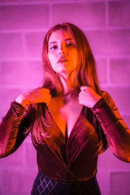 Lateks giymiş seksi kızıl kız, tuğla bir duvarda kahverengi ve siyah bir vücut giysisi, geceleri pembe ışıkla aydınlatılmış.