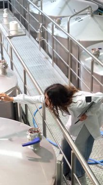 Laboratuvar önlüklü, podyum boyunca yürüyen, büyük paslanmaz çelik fermantasyon tanklarını titizlikle inceleyen ve bira üretim tesisinde bir panoya not alan bilgili bir kadın.