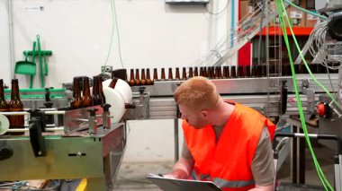 Yüksek görüş açısına sahip genç bir bira fabrikası çalışanı konveyör bandındaki boş cam şişeleri dikkatle inceliyor ve modern bir içecek fabrikasındaki bir panoya not alıyor.