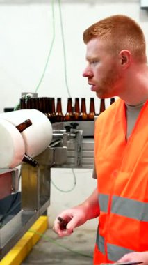 Kırmızı saçlı, güvenlik yeleği giyen ve not alan odaklı bira fabrikası çalışanı endüstriyel içecek fabrikasındaki otomatik bira şişeleme bandı sistemini dikkatle izlerken...