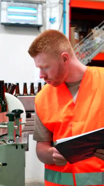 Çelik yelek giyen çalışkan fabrika işçisi bira şişeleme taşıma bandını titizlikle incelerken aynı zamanda bir not alıyor ve modern bir içecek fabrikasında kalite kontrolü sağlıyor.