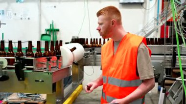 Yüksek görüş mesafesi olan bir yelek giyen genç bir erkek işçi, büyük bir endüstriyel bira fabrikasının içindeki boş cam şişe taşıyıcı sisteminde kalite kontrolü yapmak için bir pano kullanıyor.