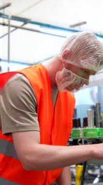 Yüksek görüş mesafesi olan bir yelek giyen erkek fabrika işletmecisi ve otomatik bira konserve ve etiketleme işlemini yöneten dokunmatik ekran paneliyle makineyi kontrol eden bir fabrika işletmecisi.