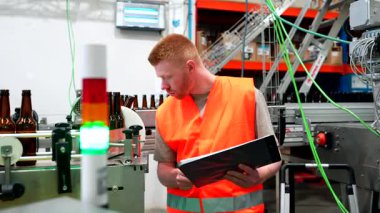 Güvenlik yeleği giymiş genç bir erkek bira fabrikası işçisi otomatik taşıma bandı sistemindeki bira şişesi üretimini dikkatle inceliyor ve kalite kontrol için bir panoya not alıyor.