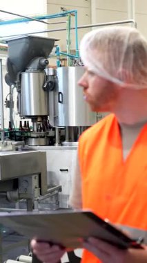 Güvenlik yeleği ve saç filesi taşıyan odaklanmış erkek fabrika işçisi, kalite kontrol için bir içecek üretim tesisinin içindeki şişe taşıma bandı ve makineleri incelerken elinde bir pano tutuyor.