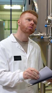 Bira imalatçısı olarak çalışan beyaz önlüklü genç adam paslanmaz çelik fermantasyon fıçılarını dikkatle inceliyor ve modern bir bira fabrikasının içindeki bir panoya not alıyor.