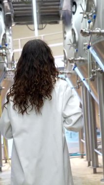 Kıvırcık saçlı, laboratuvar önlüğü giyen genç bir kadın bir bira fabrikasındaki modern mayalama ekipmanlarını incelerken büyük paslanmaz çelik fermantasyon tanklarının arasında yürüyor.