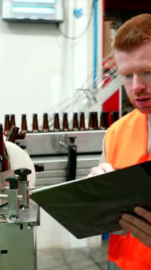 Yüksek görüş açısına sahip yeleği olan genç bira fabrikası çalışanı üretim bandındaki kahverengi cam bira şişelerini özenle inceliyor ve içecek fabrikasındaki bir panoya not alıyor.
