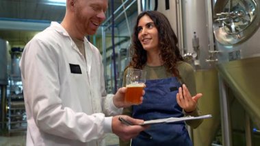 Neşeli erkek ve kadın bira üreticileri, arka planda fermantasyon tankları olan modern bir bira fabrikasındaki bir panoya not alırken yeni bir yığın zanaat birasını tartışıp tadıyorlar.
