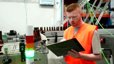 Bira fabrikasındaki taşıma bandındaki şişeleme işlemini dikkatle izleyen, kalite kontrolü yapan ve detaylı bir üretim raporu için bir panoya not alan erkek işçi.