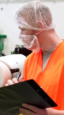 Kalite kontrol müfettişi koruyucu giysiler giyiyor. Bir yandan modern bira fabrikasındaki taşıma bandındaki şişeleme işlemini izlerken bir yandan da not alıyor. Yüksek üretim standartlarını sağlıyor.