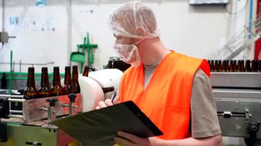 Güvenlik yeleği ve saç filesi takmış profesyonel bir erkek çalışan otomatik taşıma bandındaki boş cam şişeleri inceliyor ve modern bir içecek üretim fabrikasının içindeki bir panoya notlar yazıyor.