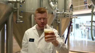 Beyaz önlüklü genç bir bira üreticisi fermantasyon tankından bir bardak taze bira dolduruyor ve tatmin olmadan önce kalitesini, rengini ve aromasını kontrol ediyor.