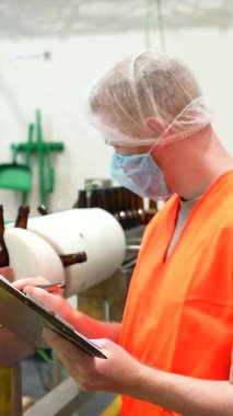 Güvenlik teçhizatı içinde çalışkan bir erkek işçi otomatik bira şişeleme taşıma bandını titizlikle inceliyor, kalite standartlarına uyması için not alıyor.