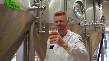 Beyaz önlüklü profesyonel bira ustası büyük paslanmaz çelik fermantasyon tankından bir ipa örneği alıyor, bira fabrikasında tadına bakmadan önce kalitesini, rengini ve aromasını kontrol ediyor.