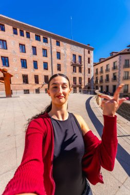 Şehir meydanında selfie çektirmek için poz veren koyu saçlı genç bir kadın, elini kaldırıyor, arkasında eski Avrupa binaları ve açık mavi bir gökyüzü var.