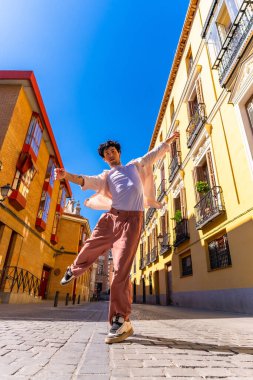 Genç adam Avrupa 'nın tarihi mimarisinin kaldırım taşı caddesinde aktif olarak poz veriyor ve dans ediyor. Parlak mavi gökyüzü altında, neşe ve enerji ifade ediyor.