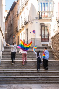 Genç yetişkinler sevinçle Avrupa 'nın bir şehrinde taştan merdivenlerden çıkıyorlar, gökkuşağı bayrakları ve çanta taşıyorlar, çeşitliliği ve Igbtq artı cemaatini bir gurur etkinliği sırasında kutluyorlar.