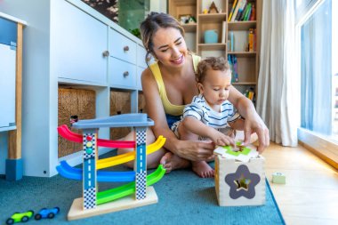 Mutlu bir anne, modern bir oturma odasındaki paspasın üzerinde oturan, şekil ayırıcı bir oyuncakla oynayan oğluna yardım ediyor.