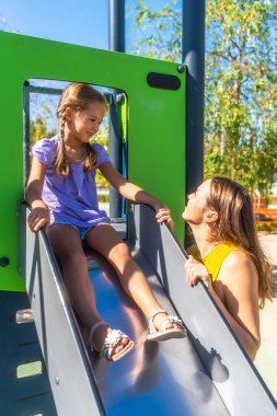 Anne, gülen kızını güneşli bir park kaydırağında cesaretlendiriyor açık hava eğlencesi sırasında neşeli bir destek, neşe ve aile bağını paylaşıyor.