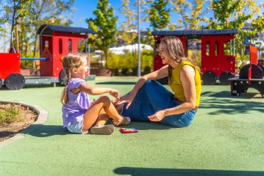 Anne ve kızı halka açık bir parkta yumuşak zeminde oturuyor güneşli bir günde mutlu bir şekilde gülüyor, birlikte oynarken bir bağ anı paylaşıyorlar.