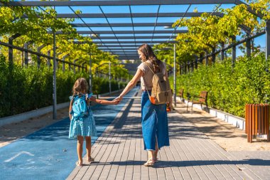 Anne ve kızı güneşli bir günde el ele tutuşarak ve sırt çantalarını taşıyarak şehir yolunda yürüyor. Bu resim aile, eğitim ve çocukluk temalarını temsil eder.
