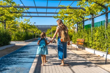 Anne ve kız, sırt çantaları takıyor, yeşil ağaçlar ve çalılarla kaplı güneşli bir patikada yürüyor, okula ya da anaokuluna giderken el ele tutuşuyorlar.