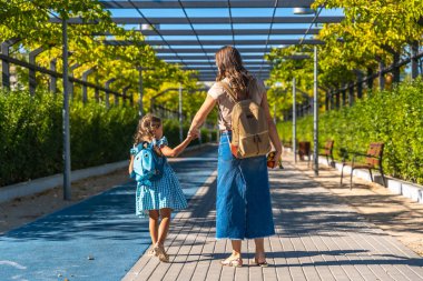 Anne ve kız el ele tutuşup asfalt bir yolda birlikte yürüyorlar. Sırt çantalarını taşıyorlar ve parlak güneşli bir günde okula gidiyorlar. Öğrenmeye hazırlar.