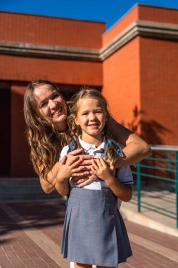 Anne ve kızı, güneşli bir günde okulda üniforması ve sırt çantasıyla bir arada dururken gülümsüyor. Destek ve ilgiyi simgeliyor.