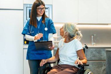 Stetoskop takan ve elinde bir dosya olan genç hemşire, evde bakım için tekerlekli sandalyede oturan yaşlı ve gülümseyen bir kadınla tedavi planını tartışıyor.