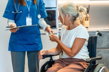 Hemşire evde tekerlekli sandalyedeki yaşlı bir kadına su toplaması için ilaç veriyor. Şefkatli bakıcı desteği, günlük tedavi ve yaşlı bakımı yardımı gösteriyor.