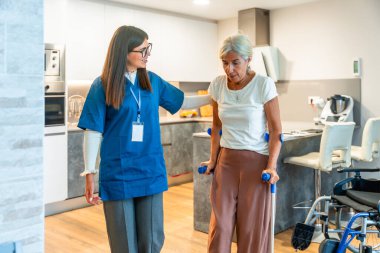 Evde bakım hemşiresi evde rehabilitasyon egzersizleri sırasında koltuk değnekleriyle yaşlı bir kadını destekliyor, hareket kabiliyetini, iyileşmeyi, bağımsızlığı ve şefkatli fizik tedaviyi teşvik ediyor