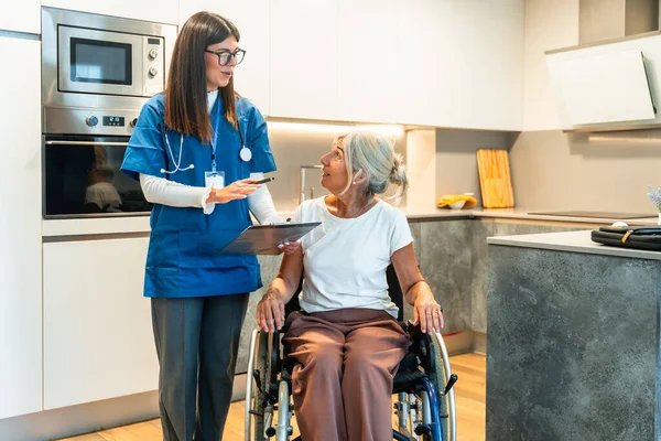 Tekerlekli sandalyede oturan yaşlı bir kadına tıbbi bilgileri açıklayan üniformalı profesyonel bir hemşire, modern bir mutfak ortamında ev sağlığı hizmetleri sunuyor.