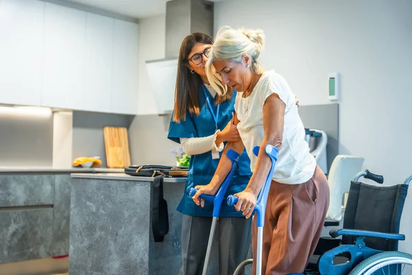 Genç bayan bakıcı, yaşlı bir kadına destek ve yardım sağlıyor. Rehabilitasyon seansında ona yardım ediyor. Mutfak ortamında bir çift koltuk değneği kullanıyor.