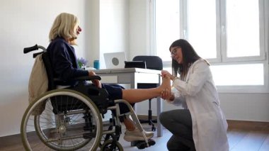 Profesyonel fizyoterapist, modern ve iyi aydınlatılmış bir klinikte rehabilitasyon sırasında tekerlekli sandalyedeki yaşlı bir kadının ayağını muayene ediyor.