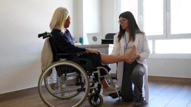 Beyaz önlüklü genç bir kadın doktor. Parlak, modern bir klinikte, tekerlekli sandalyede oturan yaşlı bir kadının ayak ve ayak bileğini muayene ediyor.