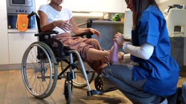 Merhametli bir bakıcı tekerlekli sandalyedeki yaşlı bir kadına fizyoterapi ve ayak masajı yapıyor. Taşınabilirlik ve ağrı kesici için evde bakım ve rehabilitasyon öneriyor.