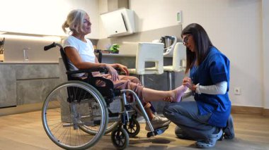 Ev ziyareti sırasında tekerlekli sandalyedeki yaşlı bir kadına fizyoterapi ve ayak masajı yapan kadın bakıcı, hasta için rehabilitasyon, hareketlilik ve ağrı kesici sunuyor.