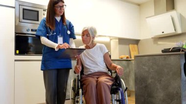 Mavi önlüklü genç bakıcı, modern bir mutfağa yaptığı ev ziyareti sırasında tekerlekli sandalyedeki yaşlı ve engelli bir hastaya tıbbi test sonuçları gösteriyor.