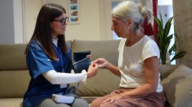 Kendini yaşlı bir kadına evde bakıcılık yapmaya adamış bir kadın hemşire. Oturma odasındaki kanepede otururken kan basıncını ölçmek için dijital monitör kullanıyor.